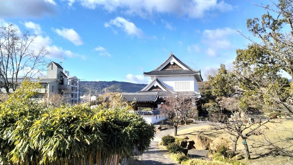 A traditional Japanese building with a tiled roof, surrounded by greenery and modern buildings in the background.