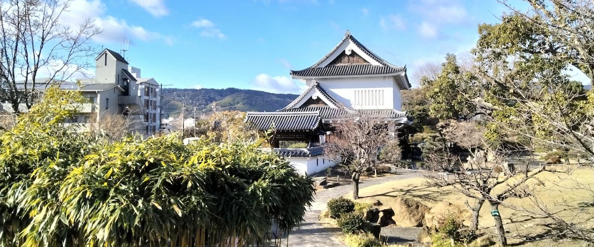 A traditional Japanese building with a tiled roof, surrounded by greenery and modern buildings in the background.