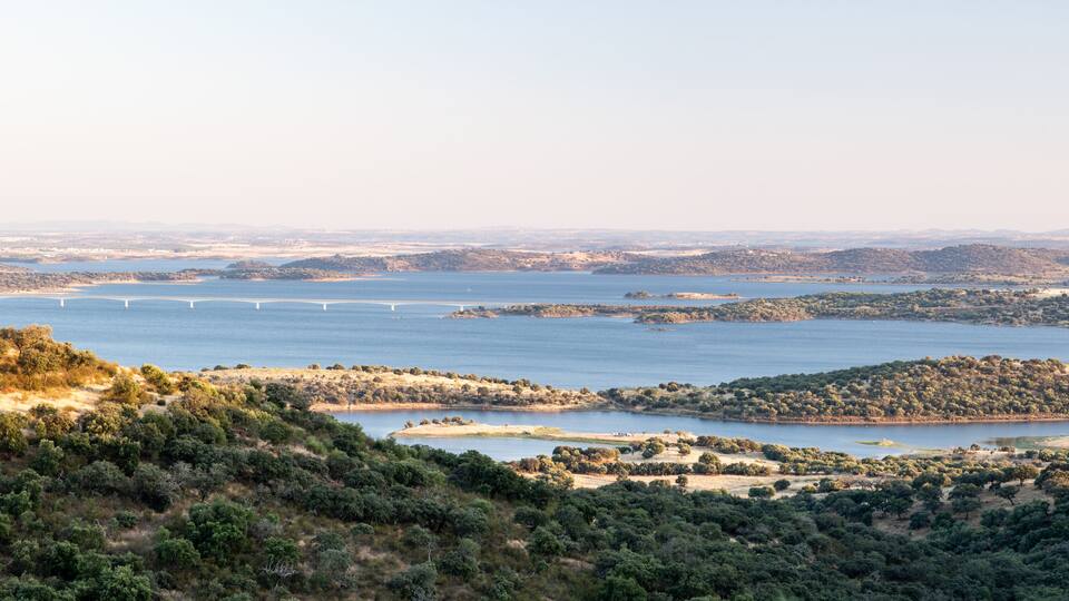 A coastal landscape with a bridge spanning a river, surrounded by hills and dense vegetation.