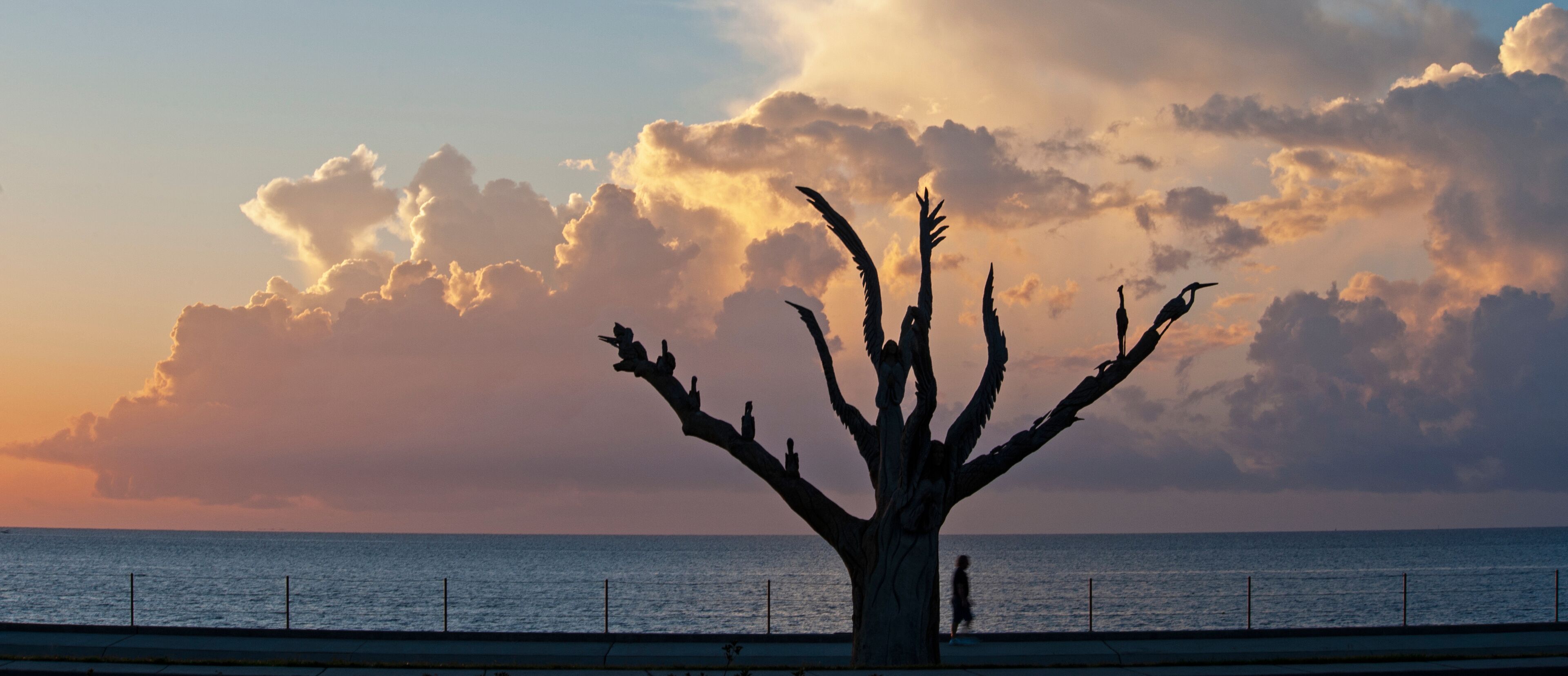 A tree sculpture with multiple branches and figures on it, set against a sunset sky with clouds and the ocean in the background.