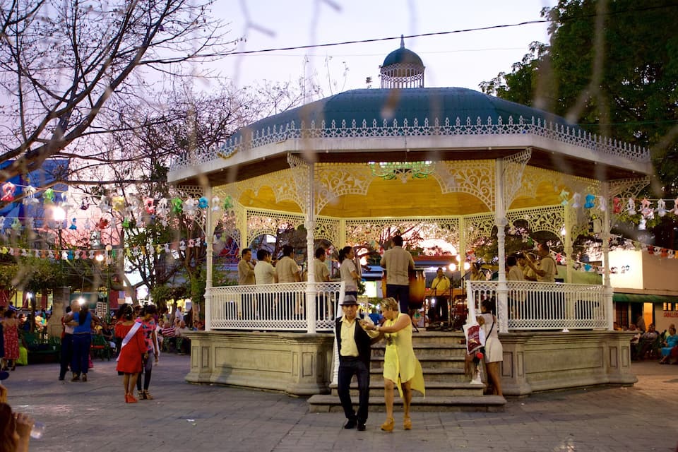 Un mirador con barandillas blancas ornamentadas y un techo abovedado azul, rodeado de gente y árboles.