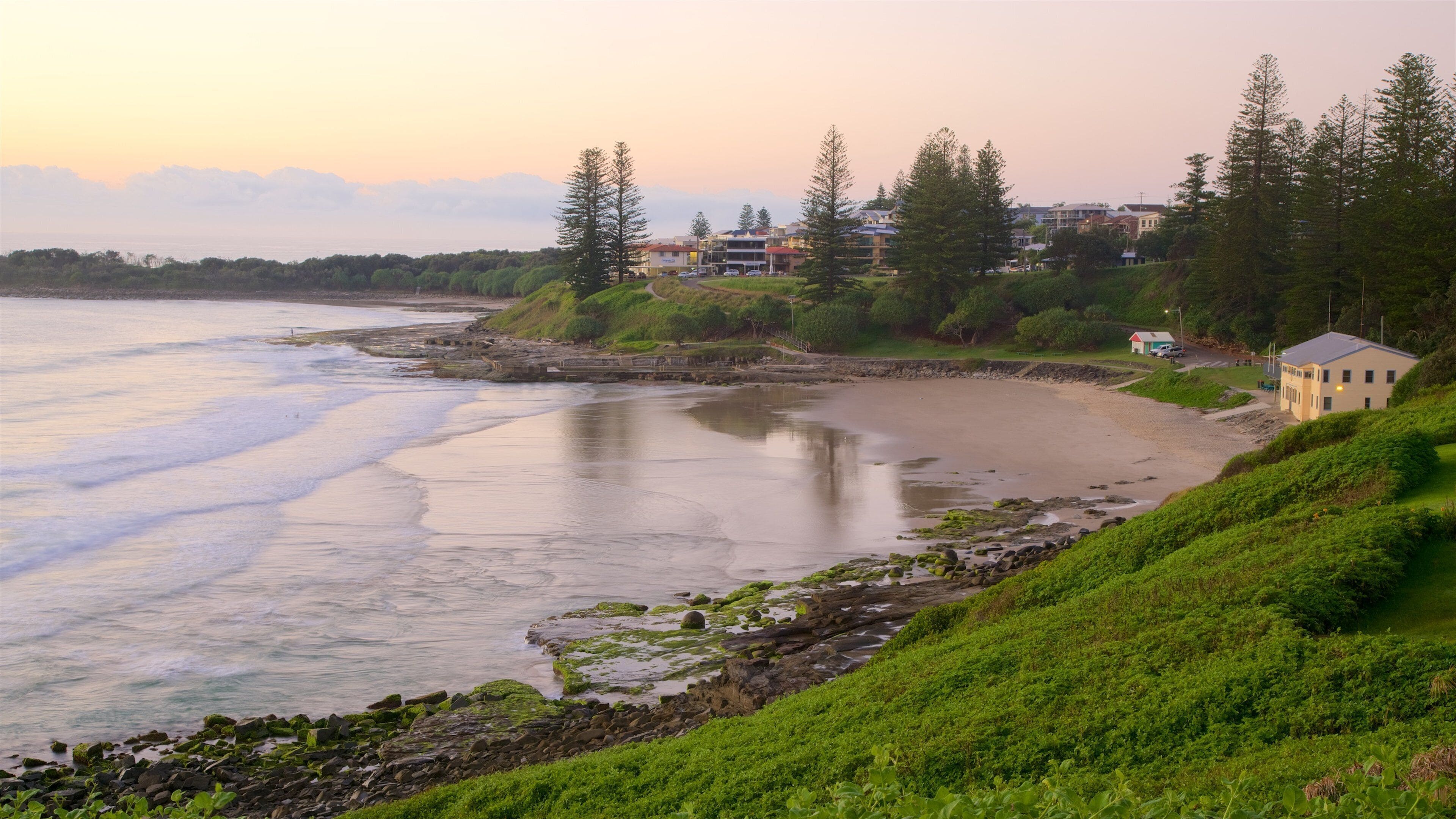 A coastal landscape with a sandy beach, rocky shoreline, green vegetation, and residential buildings in the background.