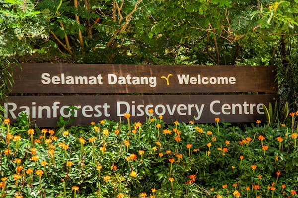Ein hölzernes Schild mit der Aufschrift 'Selamat Datang Welcome' und 'Rainforest Discovery Centre', umgeben von Grünpflanzen und orangefarbenen Blüten.