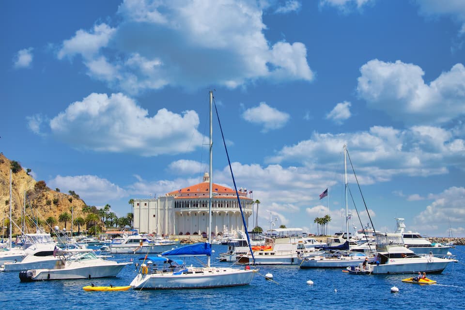 A marina with various boats docked, a large building with a red roof, and palm trees along the coastline.