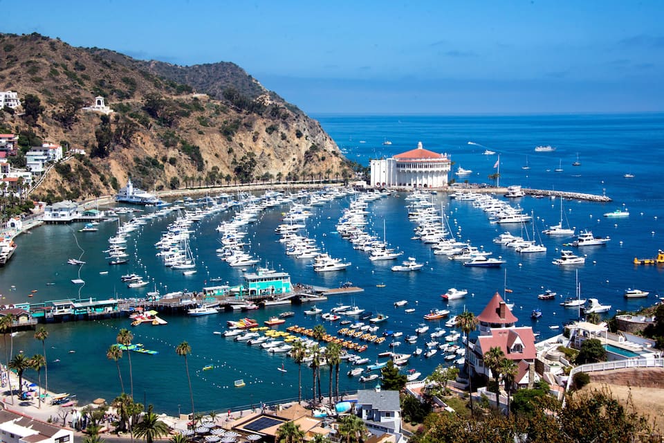 A coastal marina with numerous boats docked, a prominent white building with a red roof, and a hilly backdrop.