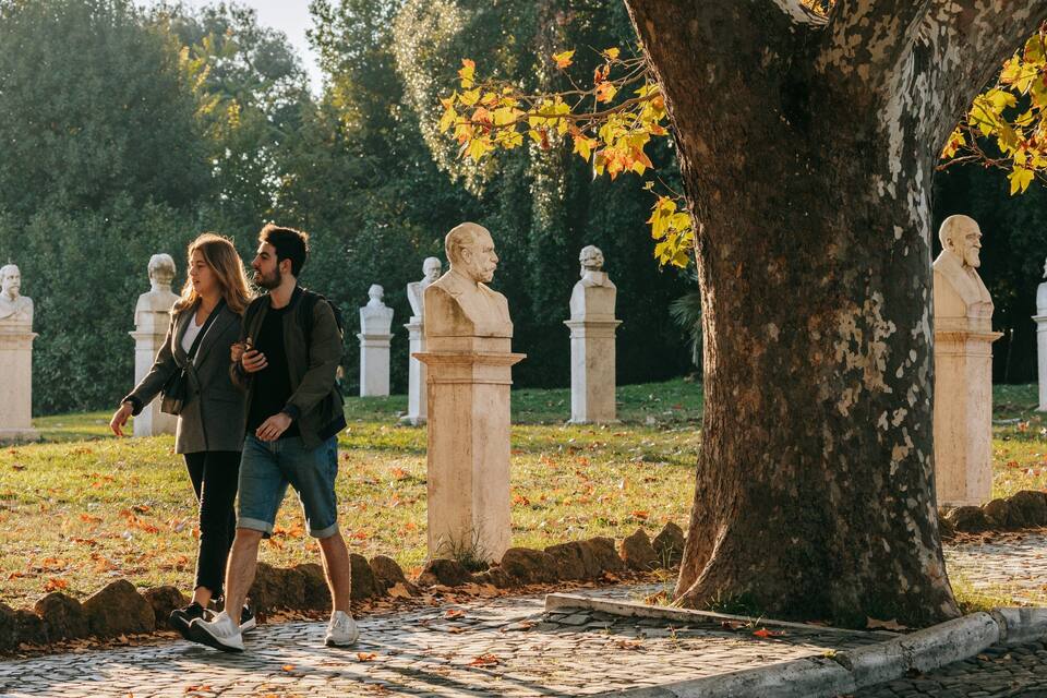 A couple walking in a park with stone sculptures and a large tree.