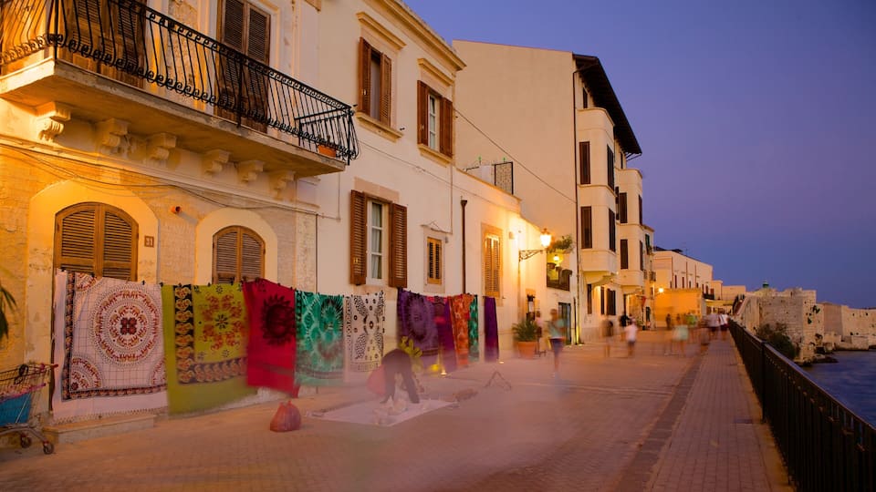 A street with colorful fabrics hanging on a building, a person washing clothes, and a historic stone wall.