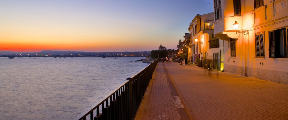 A waterfront promenade at sunset with a row of buildings and a railing along the path.