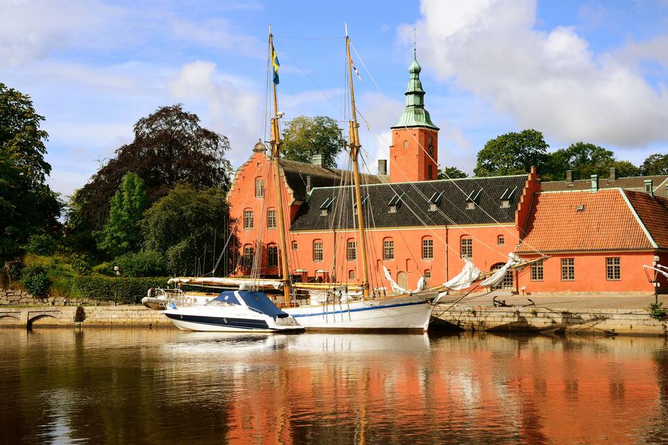 A historic building with a red facade and a green spire, a white boat docked in the foreground, and a calm river reflecting the scene.