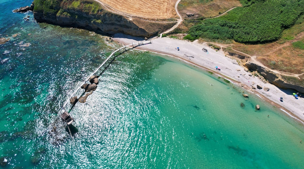 An aerial view of a coastal area with a clear beach, rocky cliffs, and a bridge connecting the land to the sea.