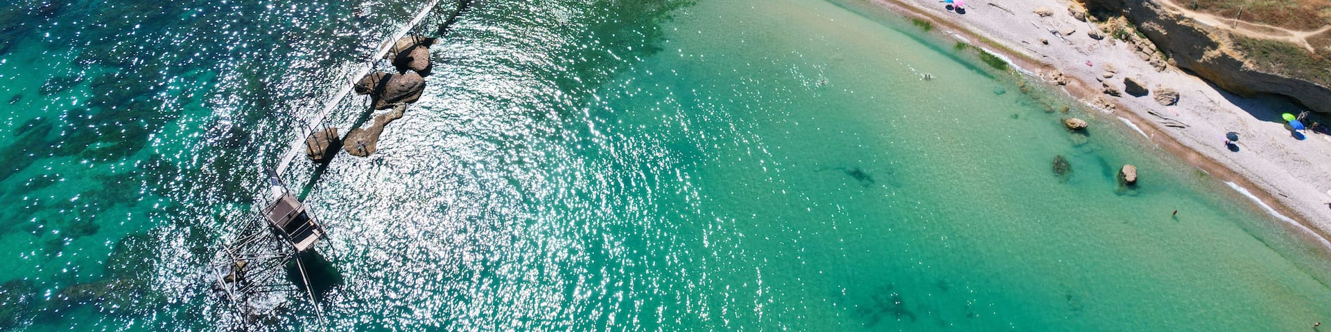 An aerial view of a coastal area with a clear beach, rocky cliffs, and a bridge connecting the land to the sea.
