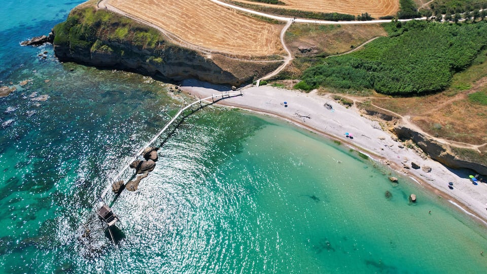 An aerial view of a coastal area with a clear beach, rocky cliffs, and a bridge connecting the land to the sea.