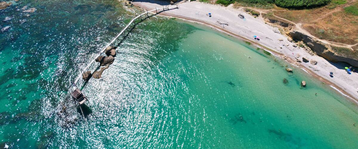 An aerial view of a coastal area with a clear beach, rocky cliffs, and a bridge connecting the land to the sea.