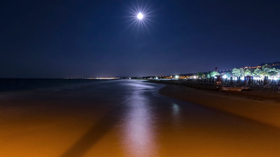 A beach at night with a bright moon and city lights in the background.