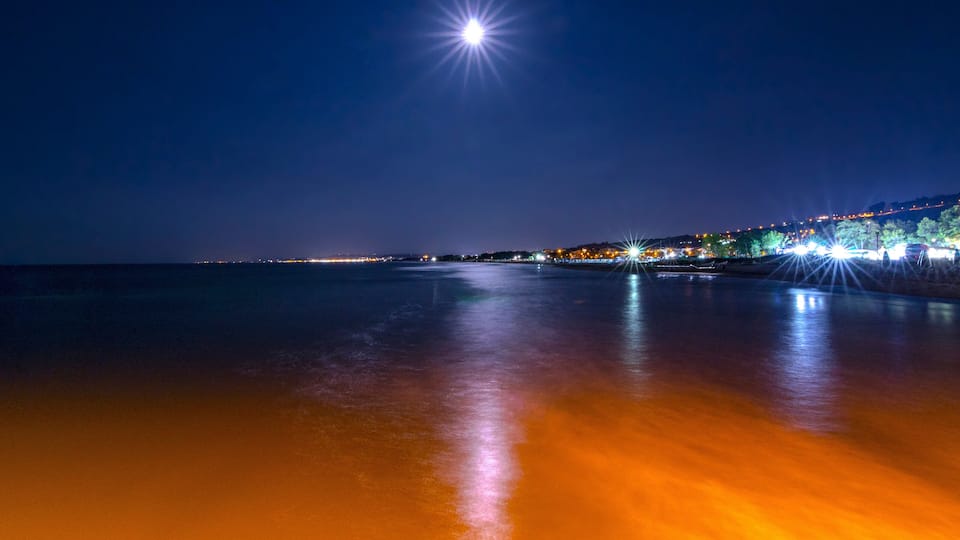 A night beach with a bright star-like light in the sky, a calm sea reflecting the lights, and a cityscape with illuminated buildings along the shore.