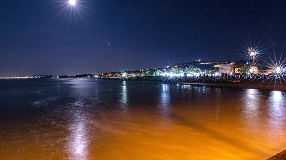 A nighttime coastal scene with a calm sea reflecting city lights, a pier, and a distant hill with buildings.