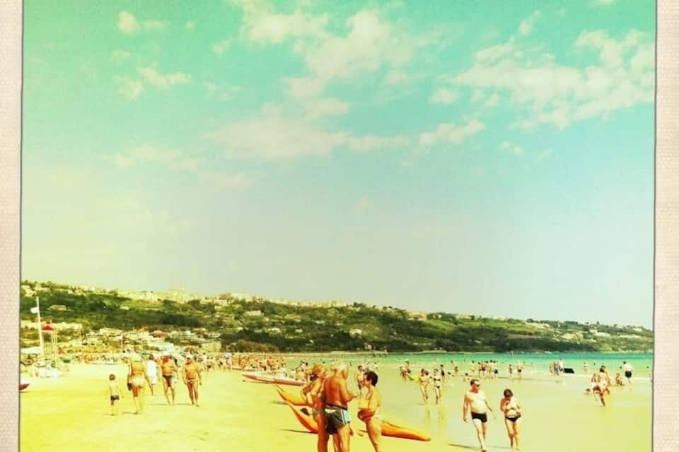 Una playa concurrida, gente caminando y llevando tablas de surf.