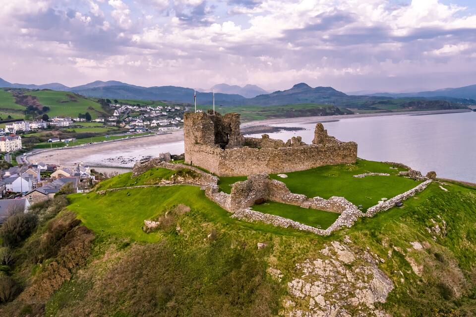 Ruinas de un castillo costero con un primer plano de césped, una playa y un pueblo al fondo.