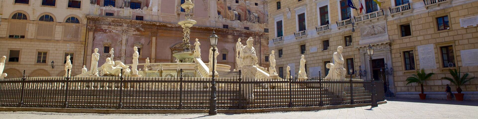A historic plaza with a fountain, classical statues, and ornate buildings.