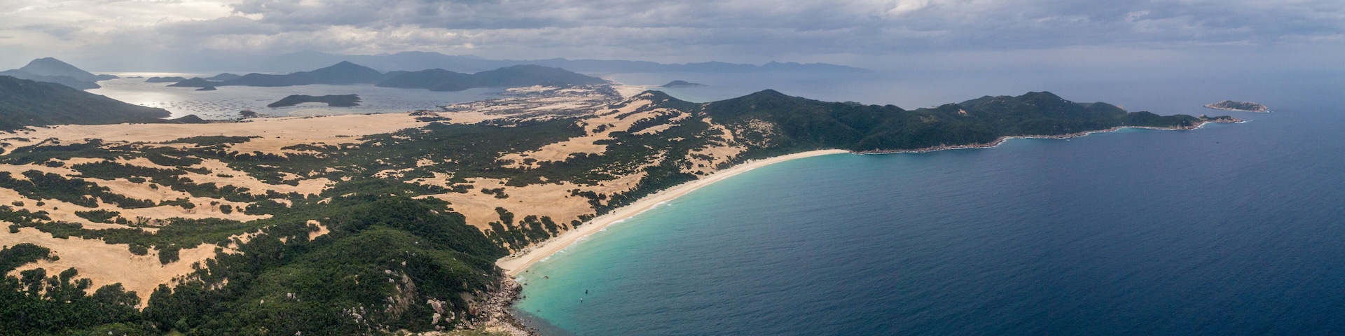 Paisagem costeira com uma praia arenosa, mar azul-turquesa e montanhas ao fundo.
