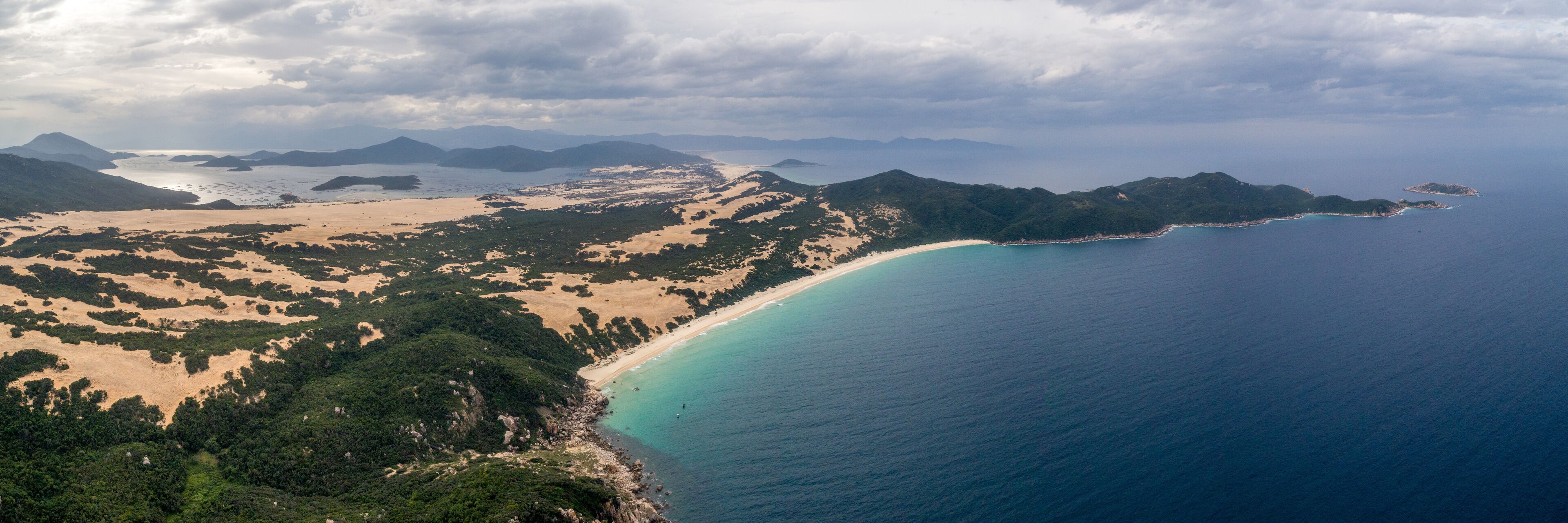 A coastal landscape with a sandy beach, a turquoise sea, and a mountainous backdrop.