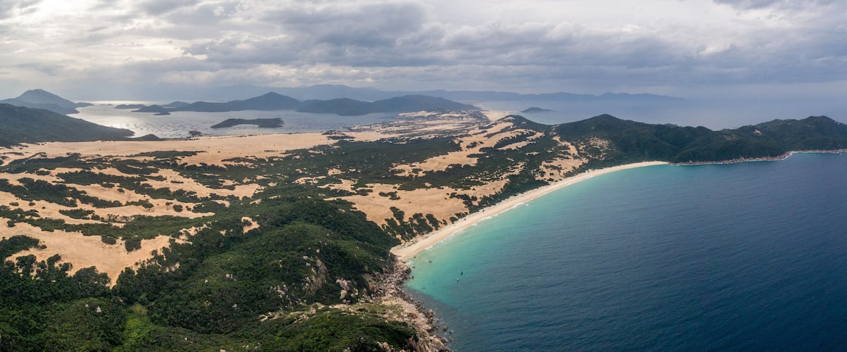 A coastal landscape with a sandy beach, a turquoise sea, and a mountainous backdrop.