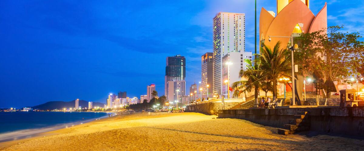 A beach with a distinctive building featuring a pointed top, palm trees, and a city skyline in the background.