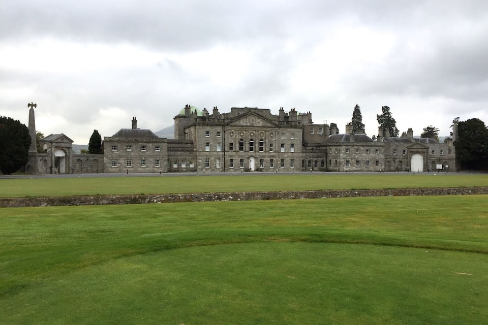 A large historic building with a green dome, a tall monument, and a well-maintained lawn in front.