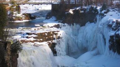 A frozen waterfall with snow-covered rocks and a bridge in the background.