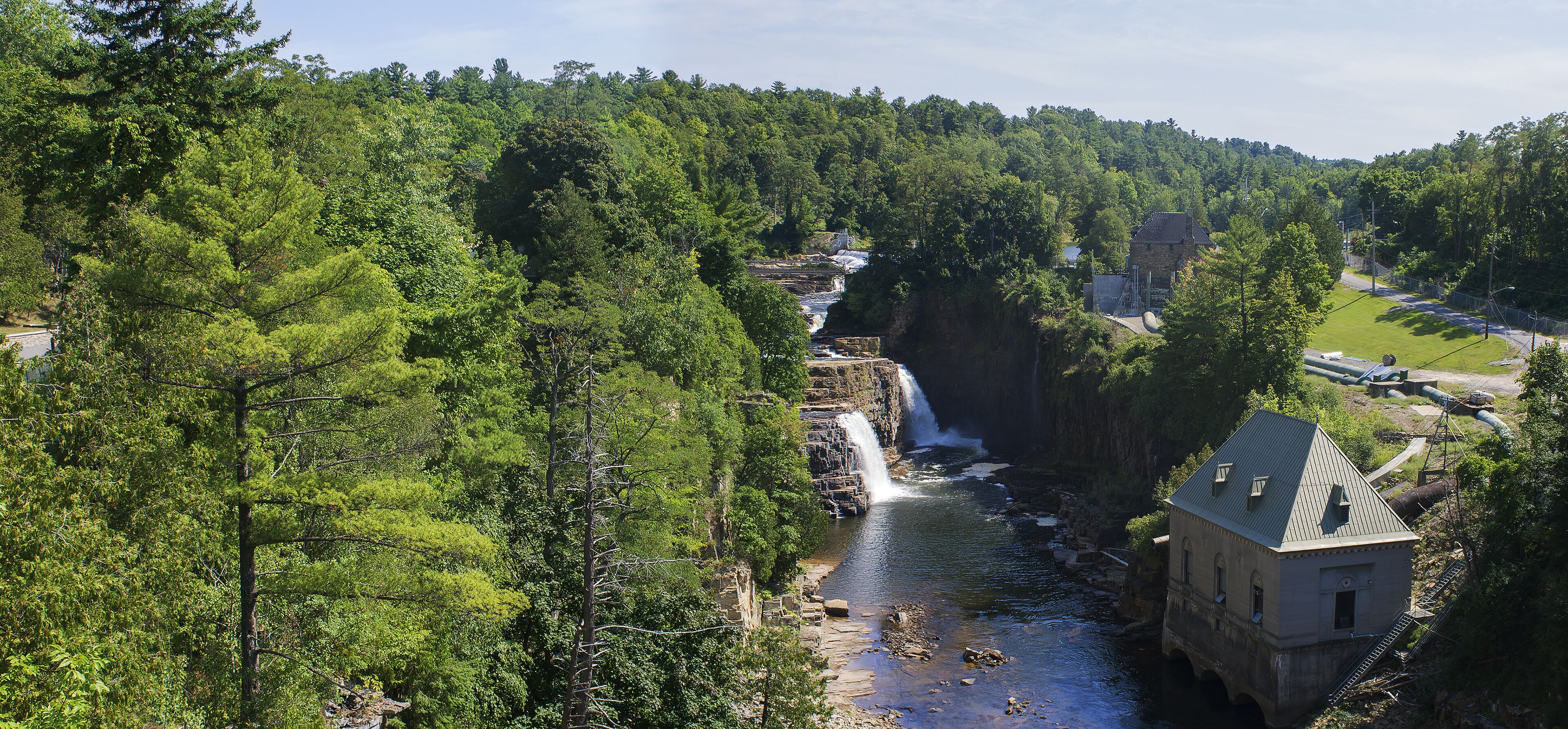 A waterfall cascading into a river surrounded by dense forest and a stone building.