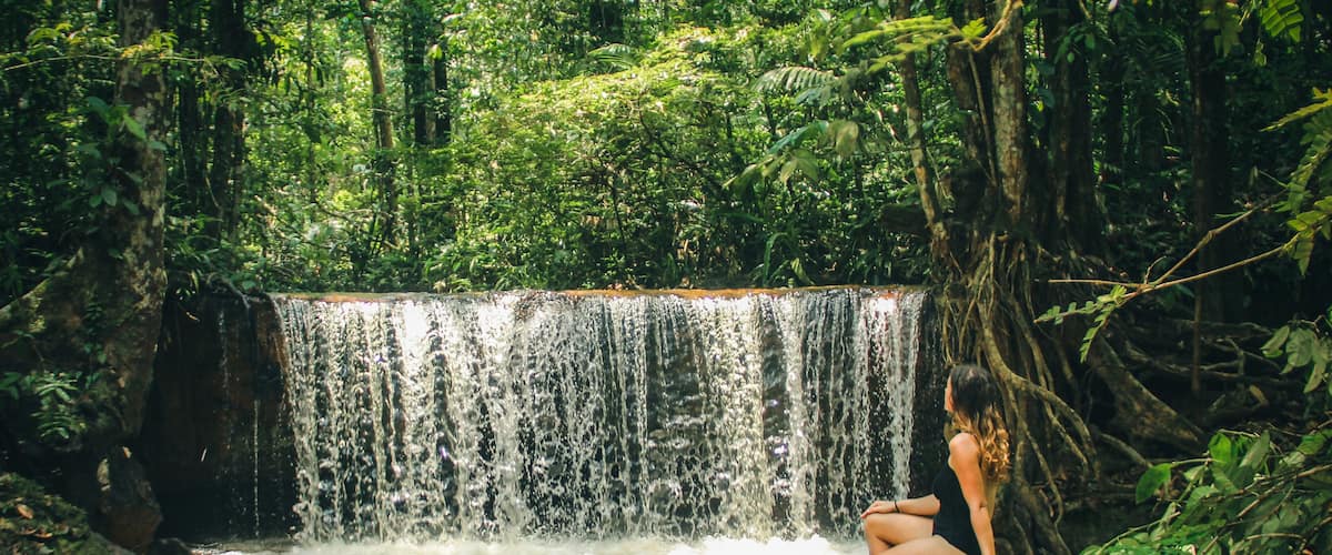 A waterfall in a forested area with a person sitting on a rock by the water.