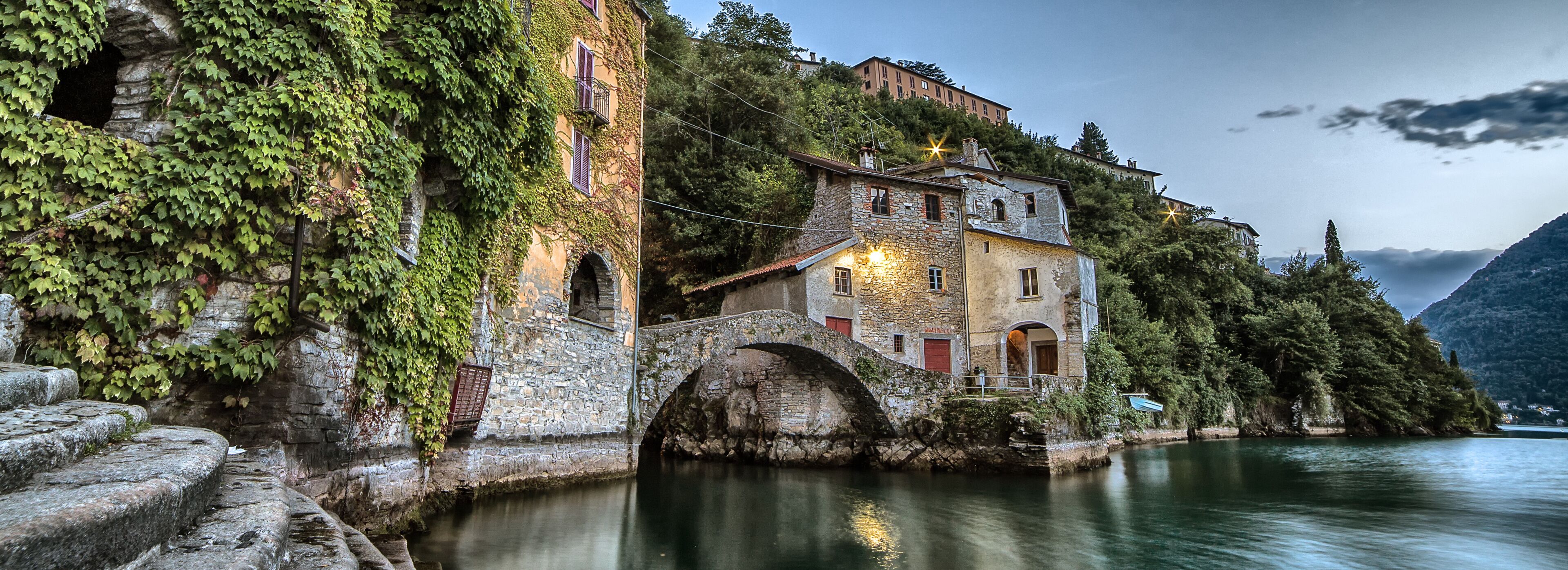 A stone bridge over a calm river, surrounded by lush greenery and historic buildings.