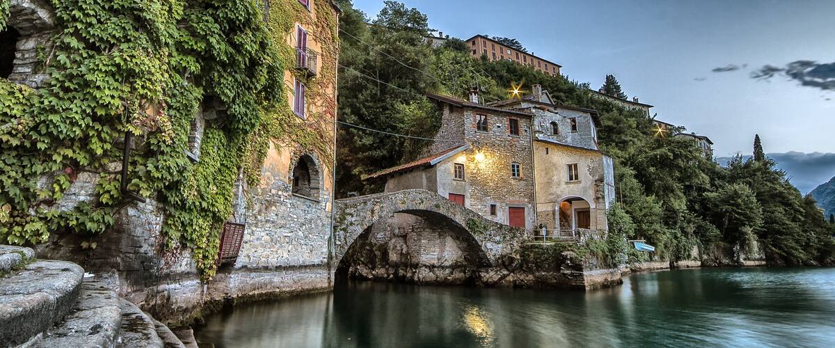 A stone bridge over a calm river, surrounded by lush greenery and historic buildings.