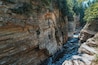 Ein Fluss, der durch eine Schlucht mit schichtartigen Felsformationen und einer Holzbrücke fließt.
