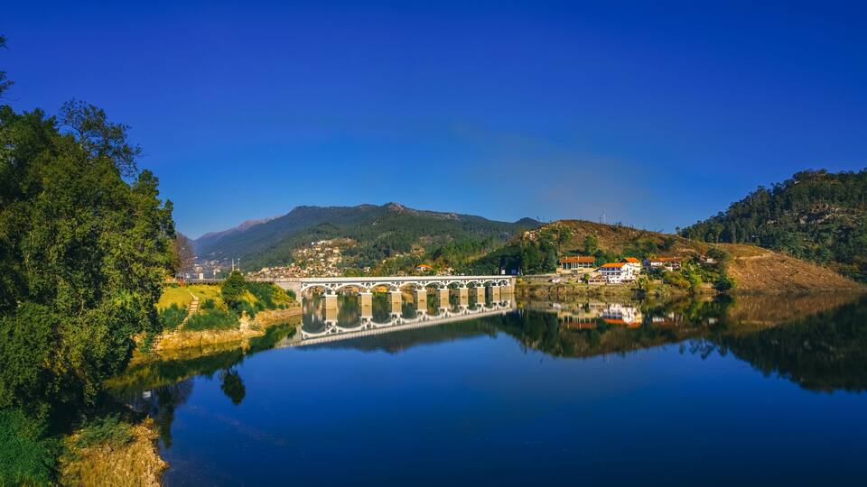 A bridge over a calm river, surrounded by lush greenery and hills.