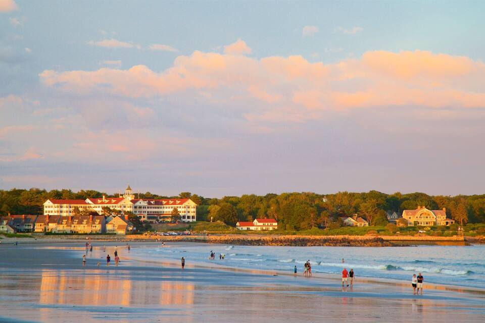 A beach with people walking and swimming, a large estate with a red roof, and a calm sea.