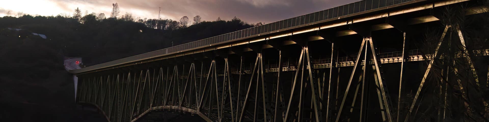 A steel bridge with a truss design, spanning over a river with a backdrop of trees and a cloudy sky.
