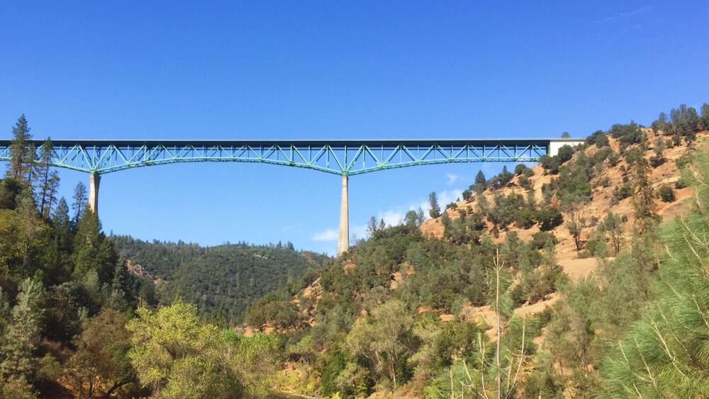A blue bridge over a river, surrounded by hills and trees.