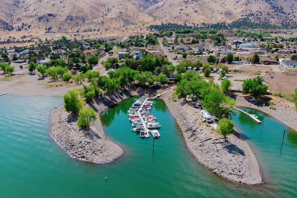 A river with boats docked on a rocky island, surrounded by a town with houses and mountains in the background.