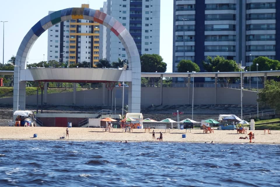 Una playa con sombrillas, una gran estructura en arco y edificios altos al fondo.