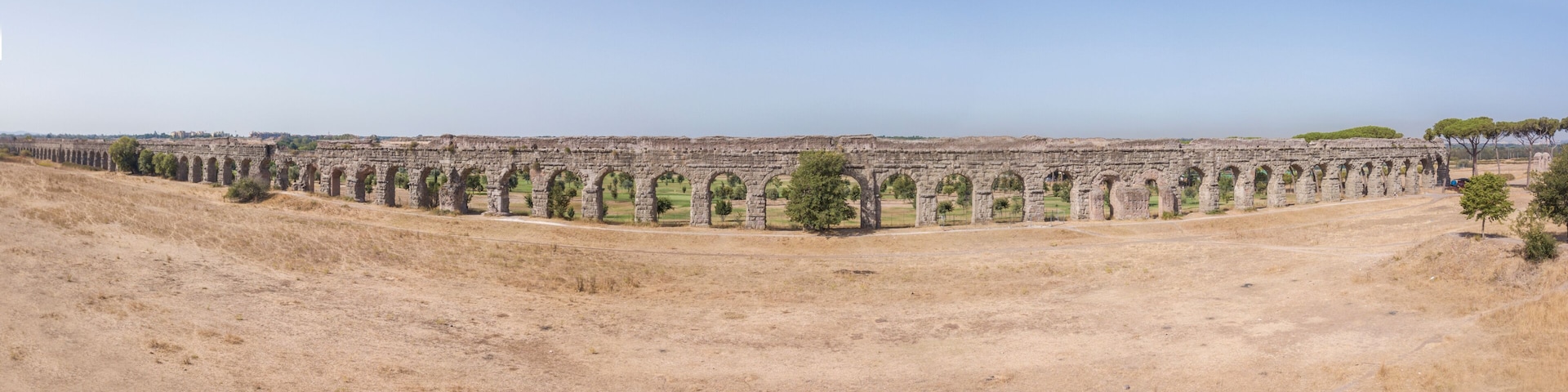 Vista aerea frontale e panoramica dei resti di un'acquedotto romano situato nel parco degli Acquedotti a Roma, vicino la fermata metro Cinecittà. In primo piano dei campi di grano.
