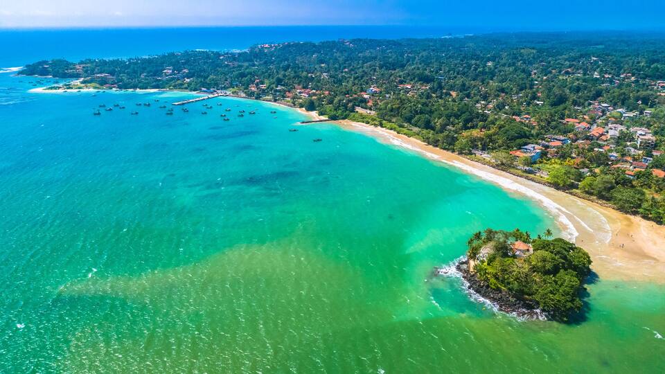 An aerial view of a coastal area with a clear turquoise sea, a sandy beach, and a densely forested island.