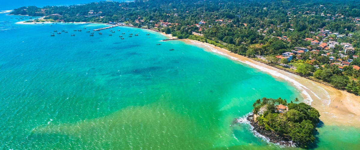 An aerial view of a coastal area with a clear turquoise sea, a sandy beach, and a densely forested island.