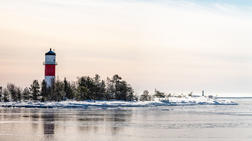 Panorama, close view of red white lighthouse in middle of frozen, snowed island at cold Baltic Sea, partly open water, thin ice reflecting day light. Blue sky, light breeze. Northern Sweden, Umea