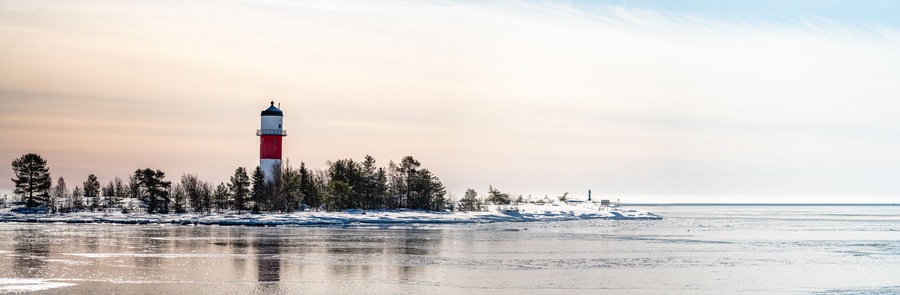 Panorama, close view of red white lighthouse in middle of frozen, snowed island at cold Baltic Sea, partly open water, thin ice reflecting day light. Blue sky, light breeze. Northern Sweden, Umea