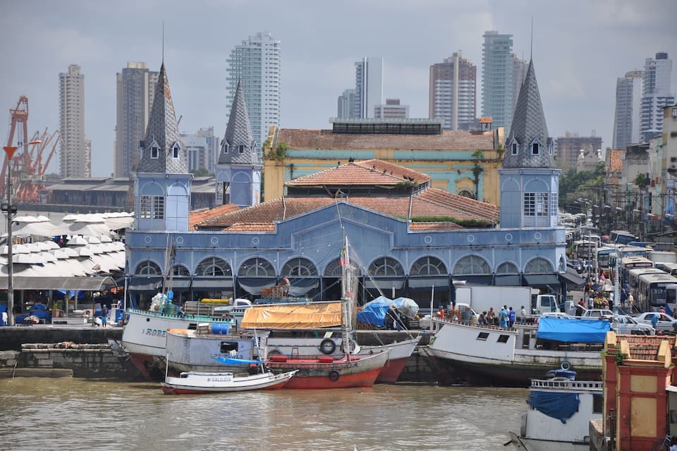 A harbor with boats docked, a large building with a distinctive roof, and a city skyline in the background.