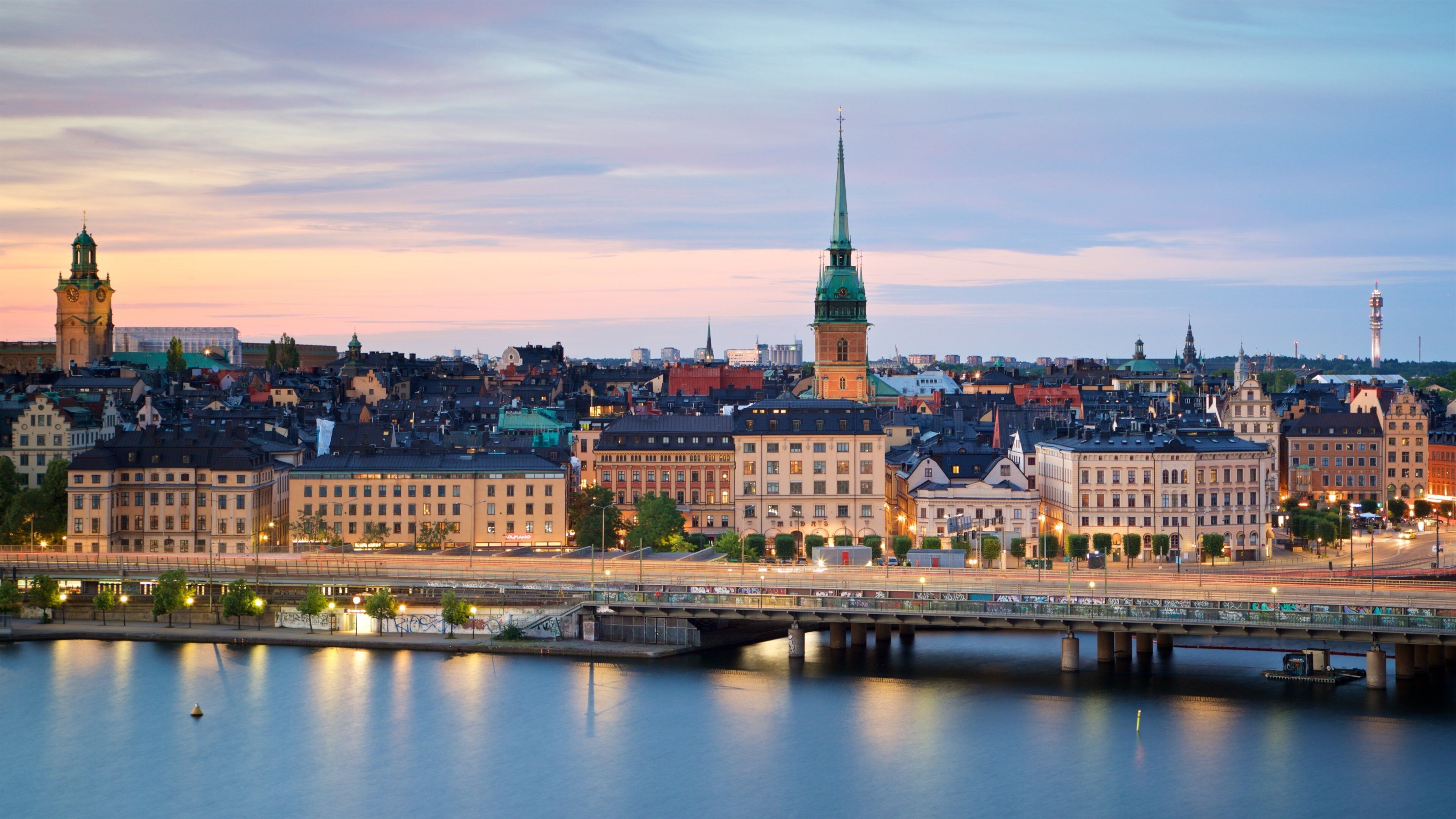 A cityscape with a prominent church spire, a clock tower, and a bridge over a calm river.