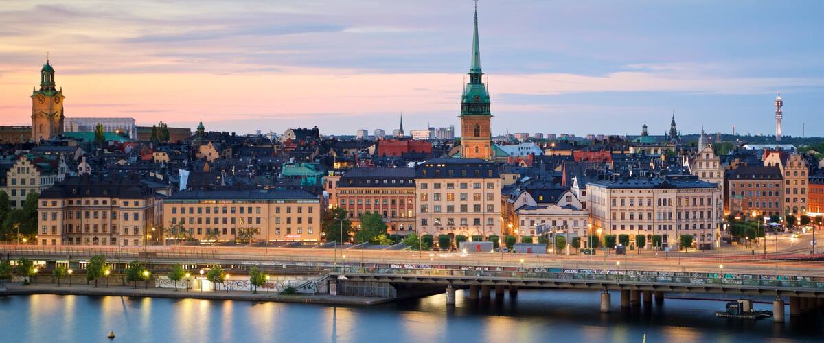 A cityscape with a prominent church spire, a clock tower, and a bridge over a calm river.
