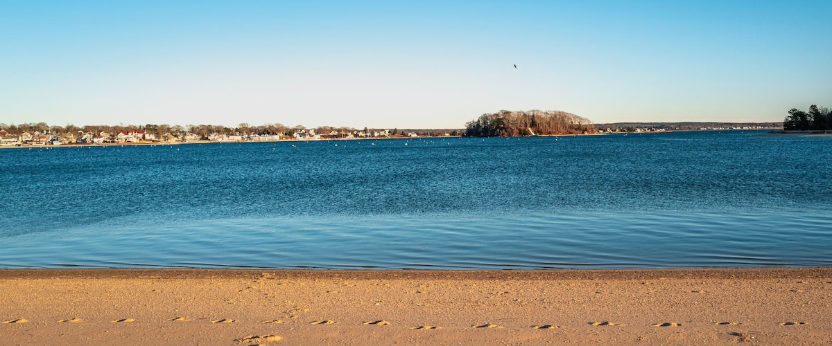 A calm lake with a sandy beach, a small island, and a distant shoreline with buildings.