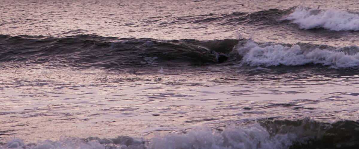 A pattern of three waves making their way to the beach and crashing on the shallow water. Each wave has a whitecap. The ocean is beneath a cloud filled sky lit by the rising sun with pink and orange.
